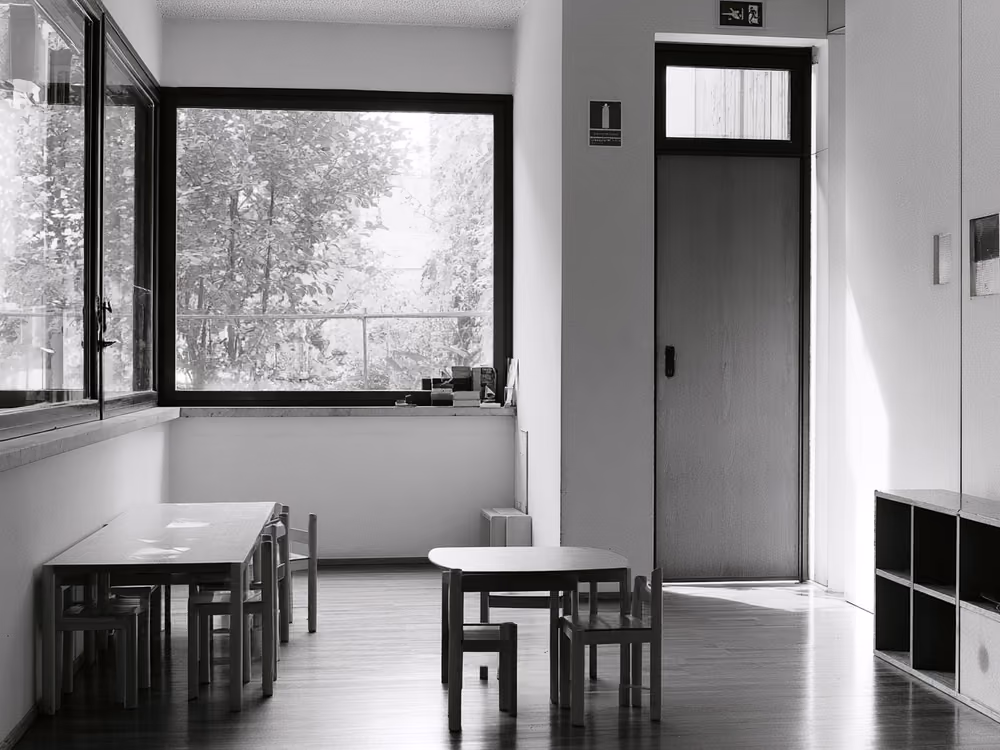 Aula dell'asilo nido con finestre basse, pavimento in legno chiaro e l'uscita di sicurezza segnalata accanto alla porta